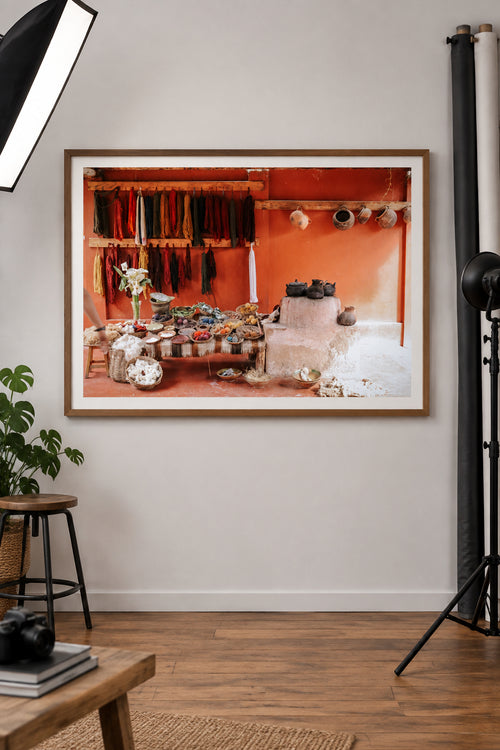 A rustic interior scene in South America with a table covered in colorful, naturally dyed yarns, wool bundles, and small bowls, set against a terracotta wall with hanging colorful textiles and clay pots.