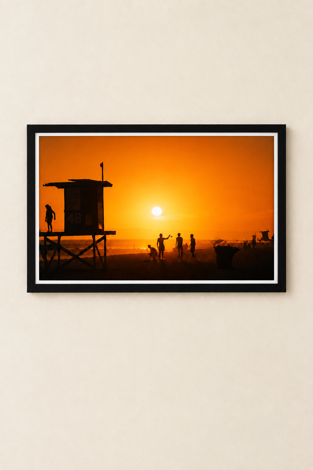 Golden orange beach sunset with silhouetted lifeguard tower and beachgoers, in a black frame with a thin white border